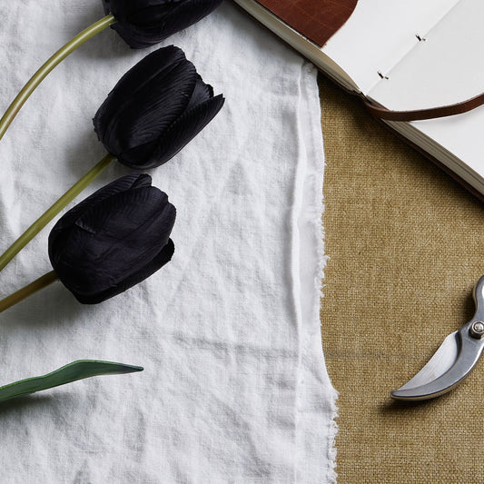 Tall black artificial tulip stems with sleek petals and green leaves, arranged on white fabric beside a notebook and scissors.
