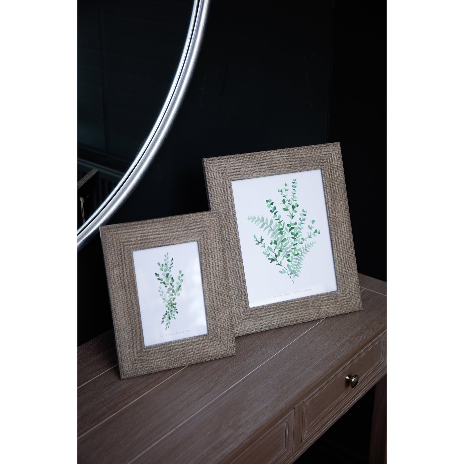 Woven photo frame in grey-washed finish, featuring intricate patterns, showcasing botanical prints on a wooden table with a round mirror.