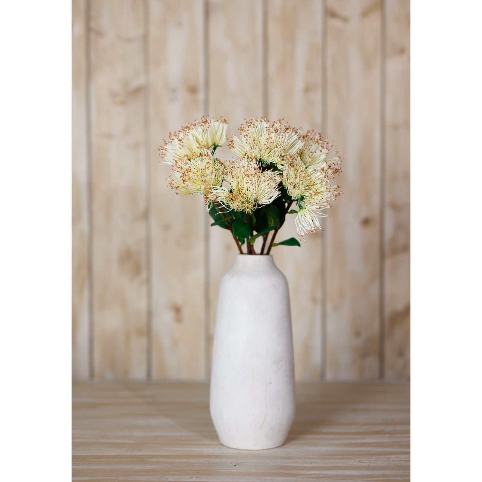 Luxurious faux Protea Nitida stem with large creamy-white blooms and lush green leaves in a simple white vase, set against a wooden background.