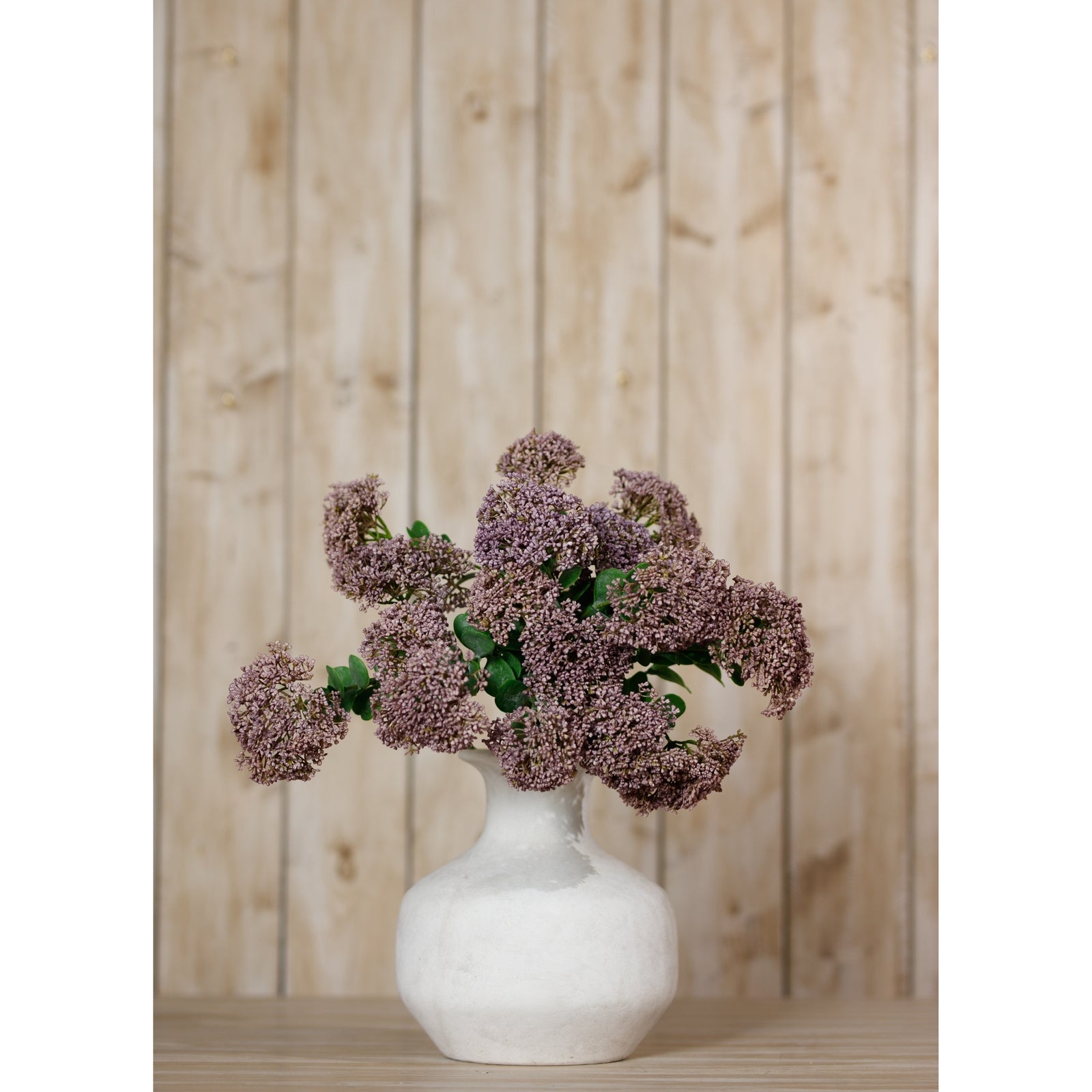 Elegant purple Sedum blooms in a white vase, complemented by lush greenery, against a light wooden backdrop.