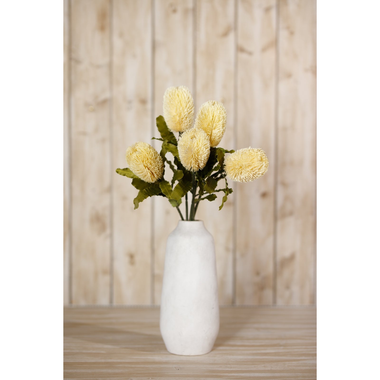 Yellow Banksia Serrata stems in a textured white vase, surrounded by lush green leaves, set against a rustic wooden backdrop.