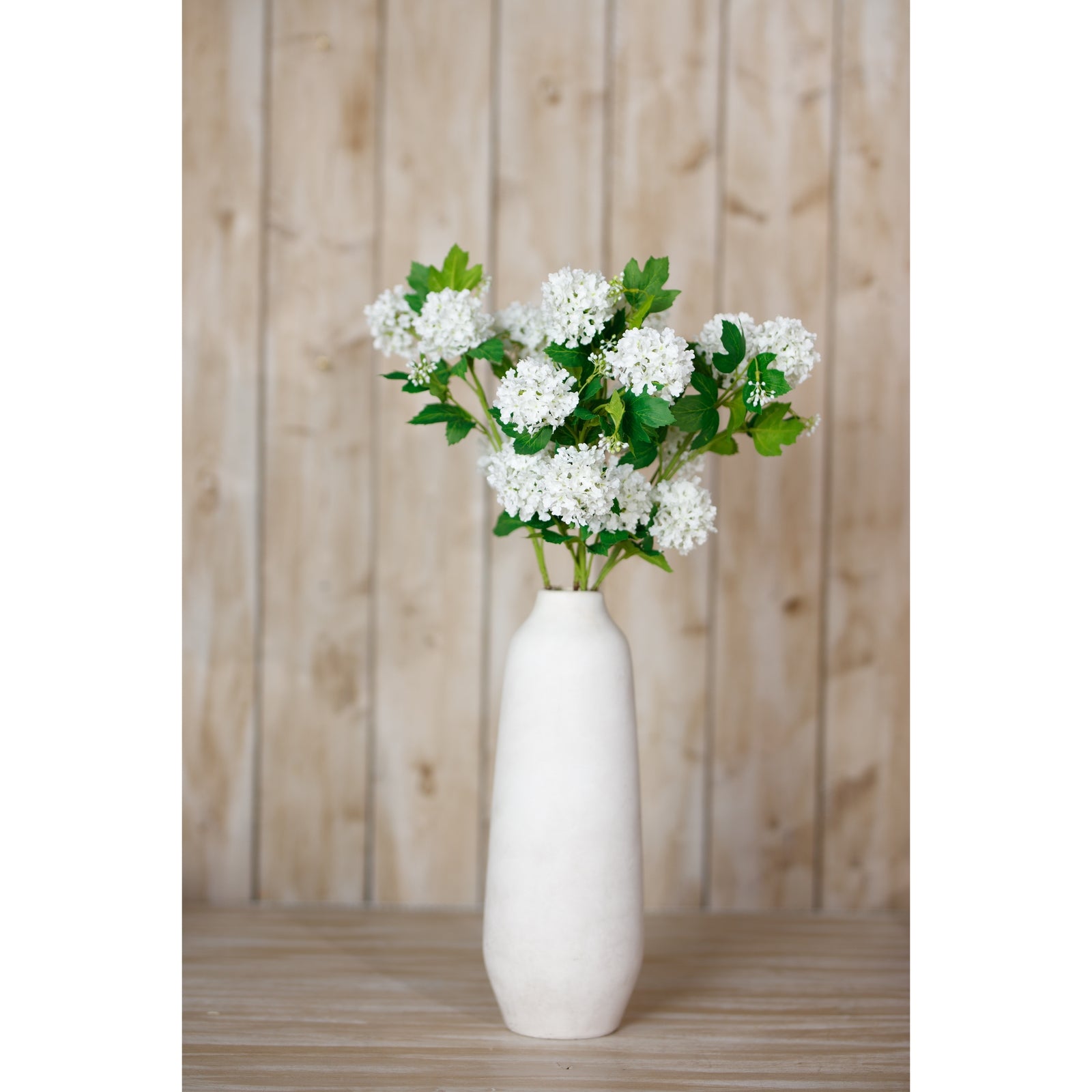 Lifelike Cream Snowball Viburnum spray with white blooms and lush green foliage in a simple, elegant vase against a rustic backdrop.