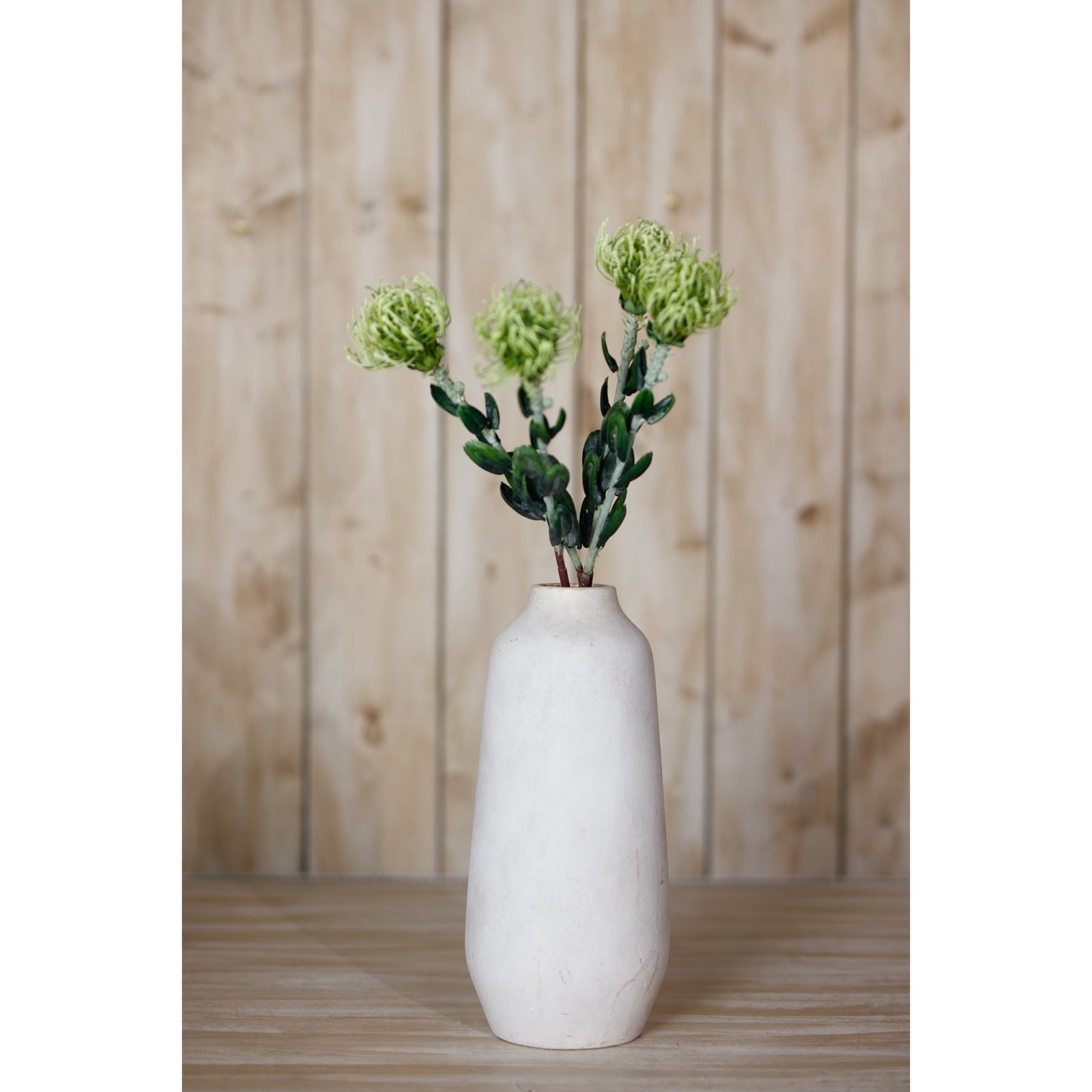 Green pincushion protea spray with lifelike petals, displayed in a simple white vase, against a natural wooden backdrop.