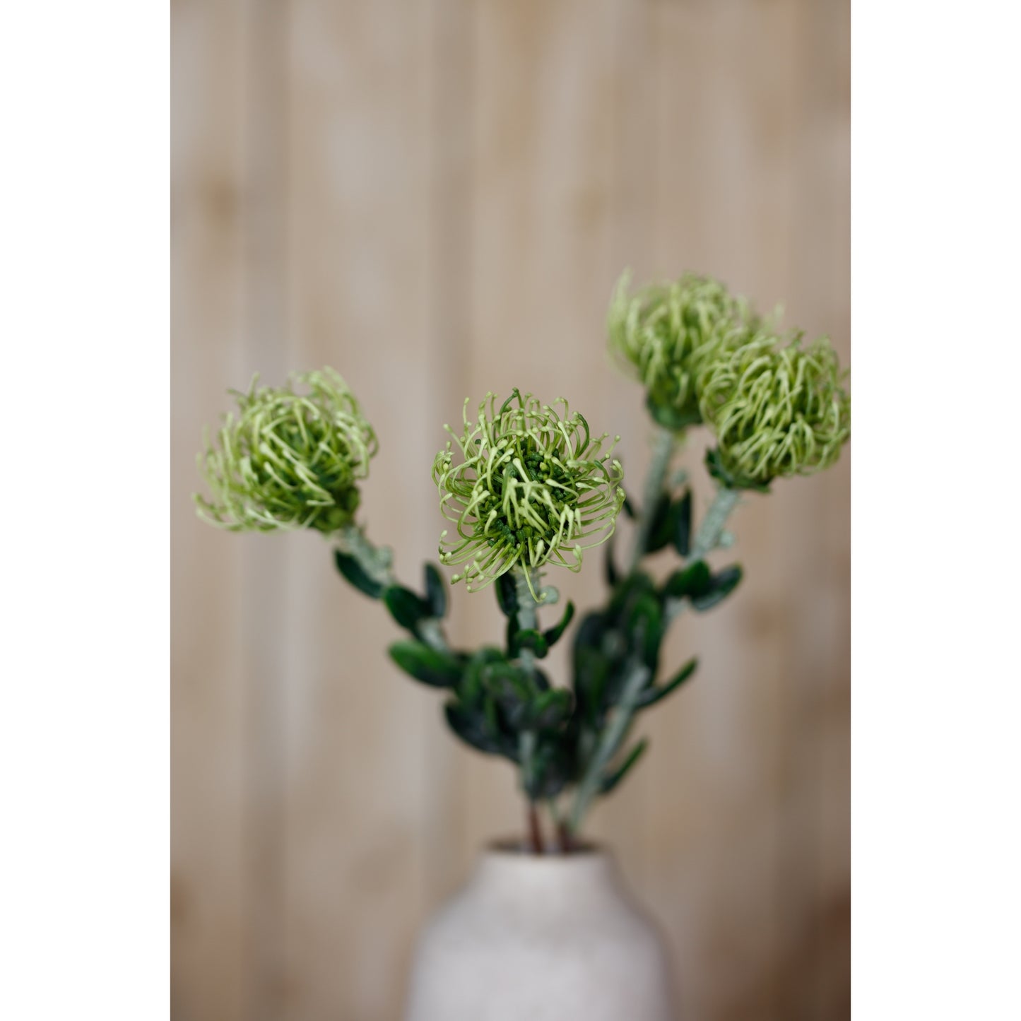 Lifelike green pincushion protea spray with intricate petals and lush leaves, displayed in a neutral vase against a warm background.