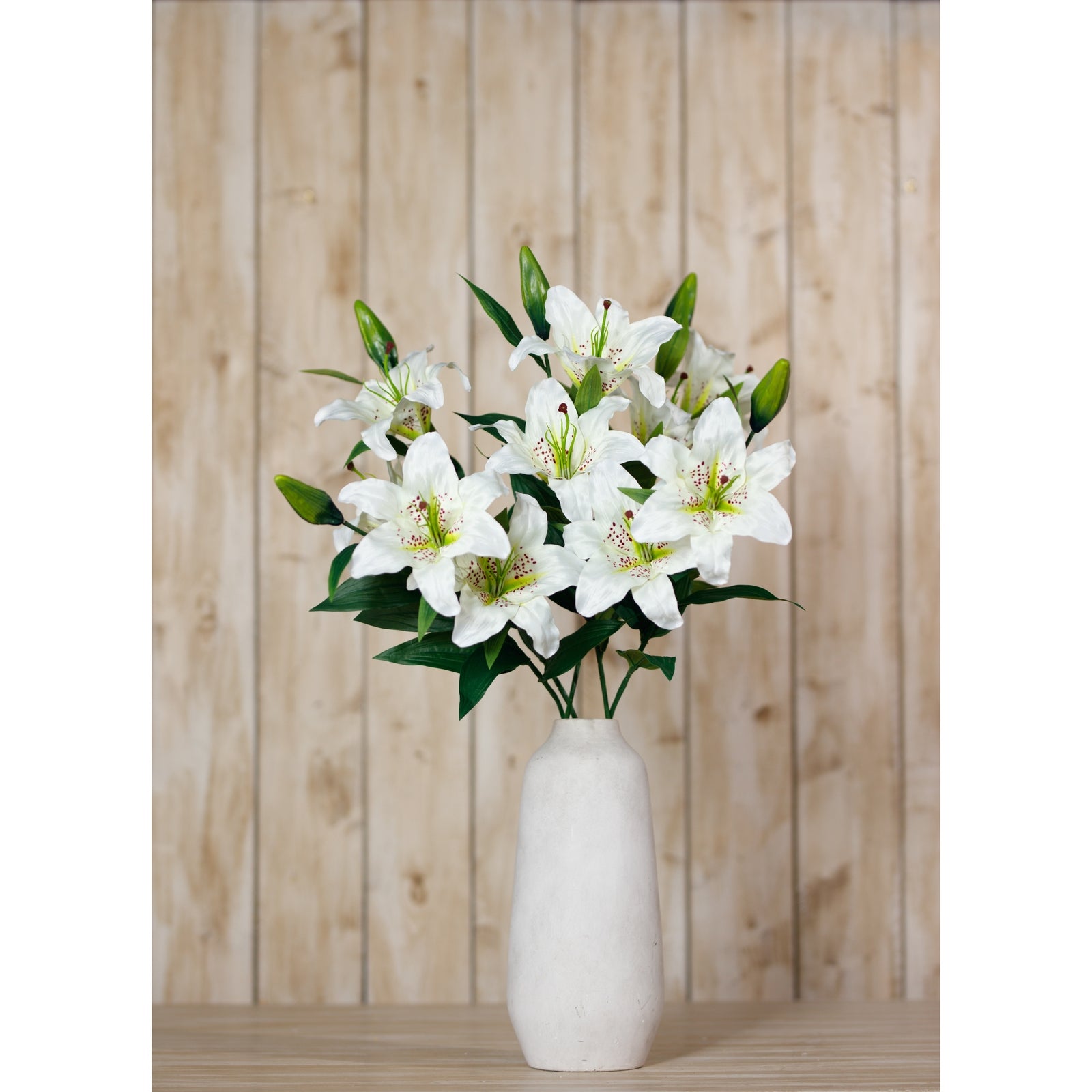Lifelike white Madonna lily spray with delicate blooms and rich green foliage in a smooth, pale vase against a wooden backdrop.