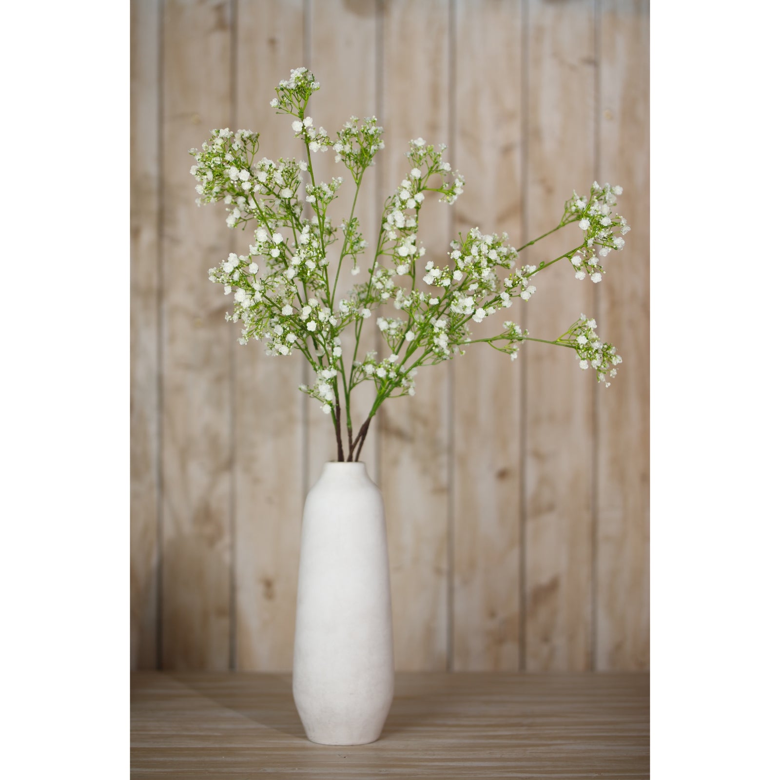 Cream Gypsophila spray features delicate clusters of tiny white flowers on green stems, elegantly arranged in a tall, soft gray vase against a wooden backdrop.