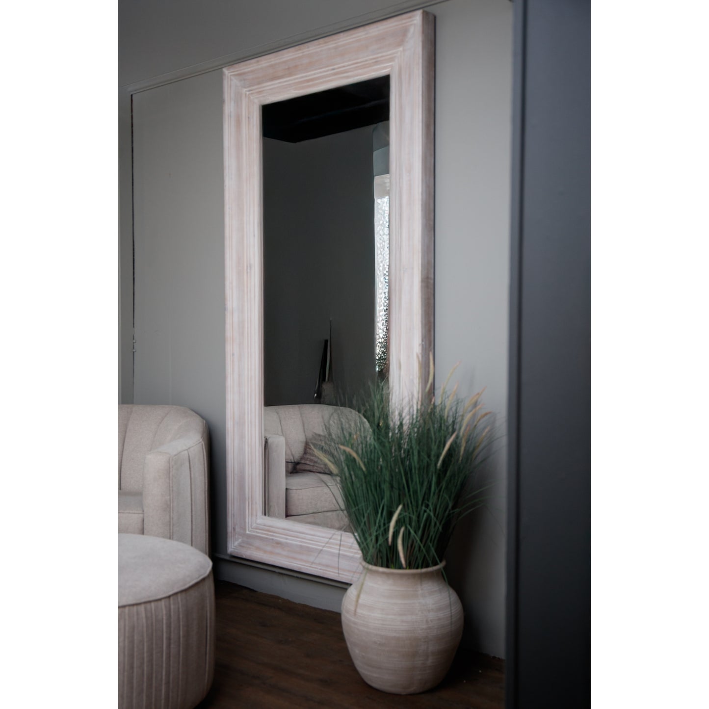 Large white-washed mirror with a weathered wood frame, reflecting a cozy interior, complemented by a stylish plant and soft furnishings.