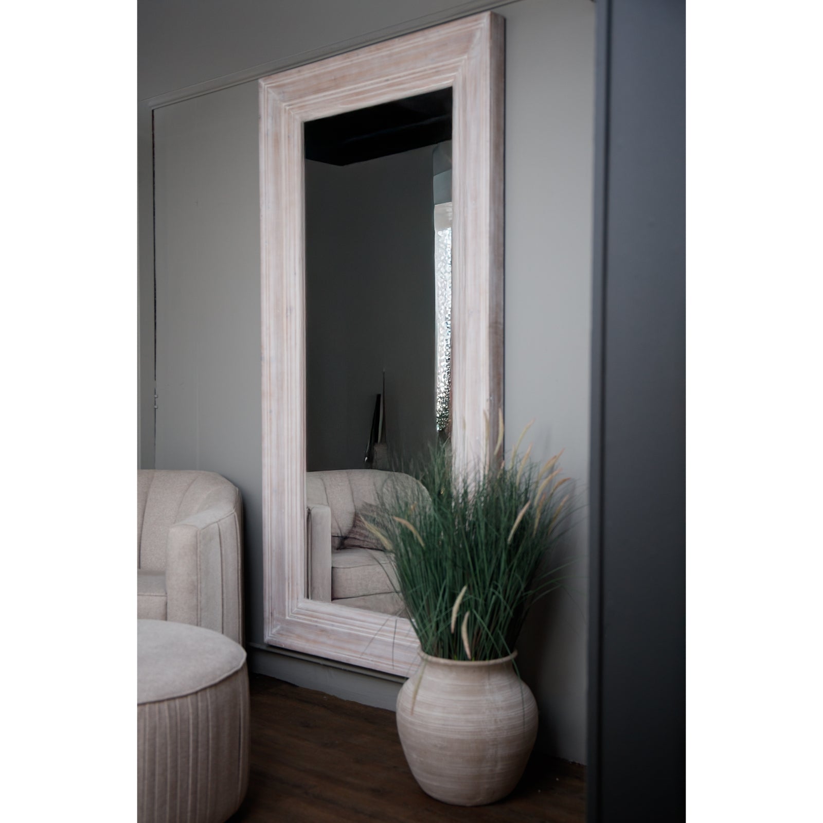Large white-washed mirror with a weathered wood frame, reflecting a cozy interior, complemented by a stylish plant and soft furnishings.