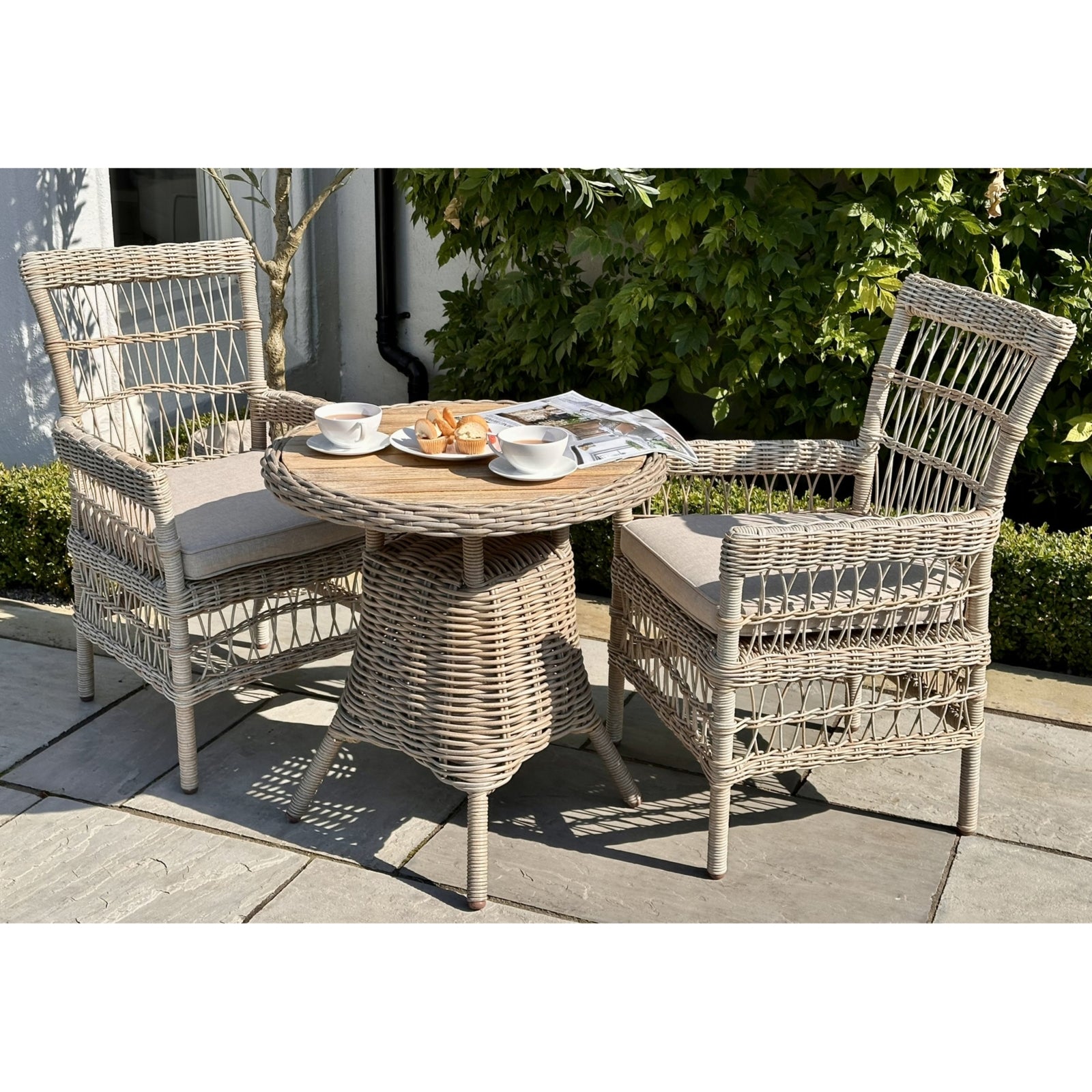 Light gray rattan outdoor dining chairs with removable beige cushions, paired with a round wicker table, set in a lush garden.