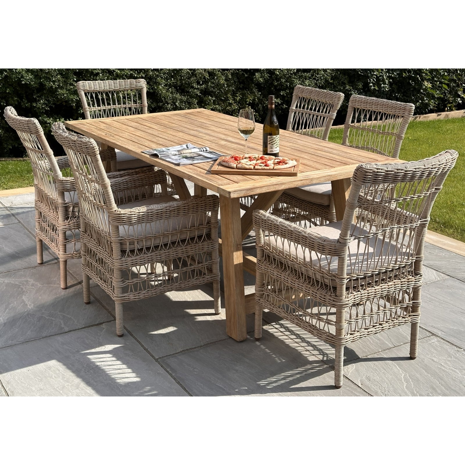Large outdoor dining table made of weather-resistant acacia wood, surrounded by elegant woven chairs, set with a wine bottle and food.