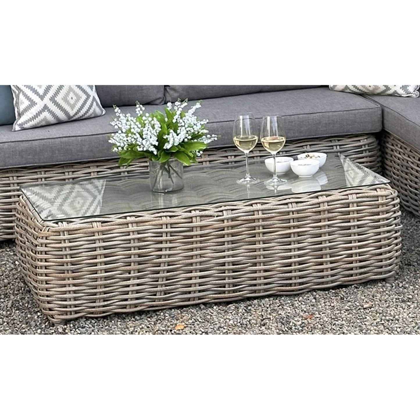 Large outdoor corner seating set in gray wicker, featuring a glass-topped table, decorative flowers, and wine glasses.