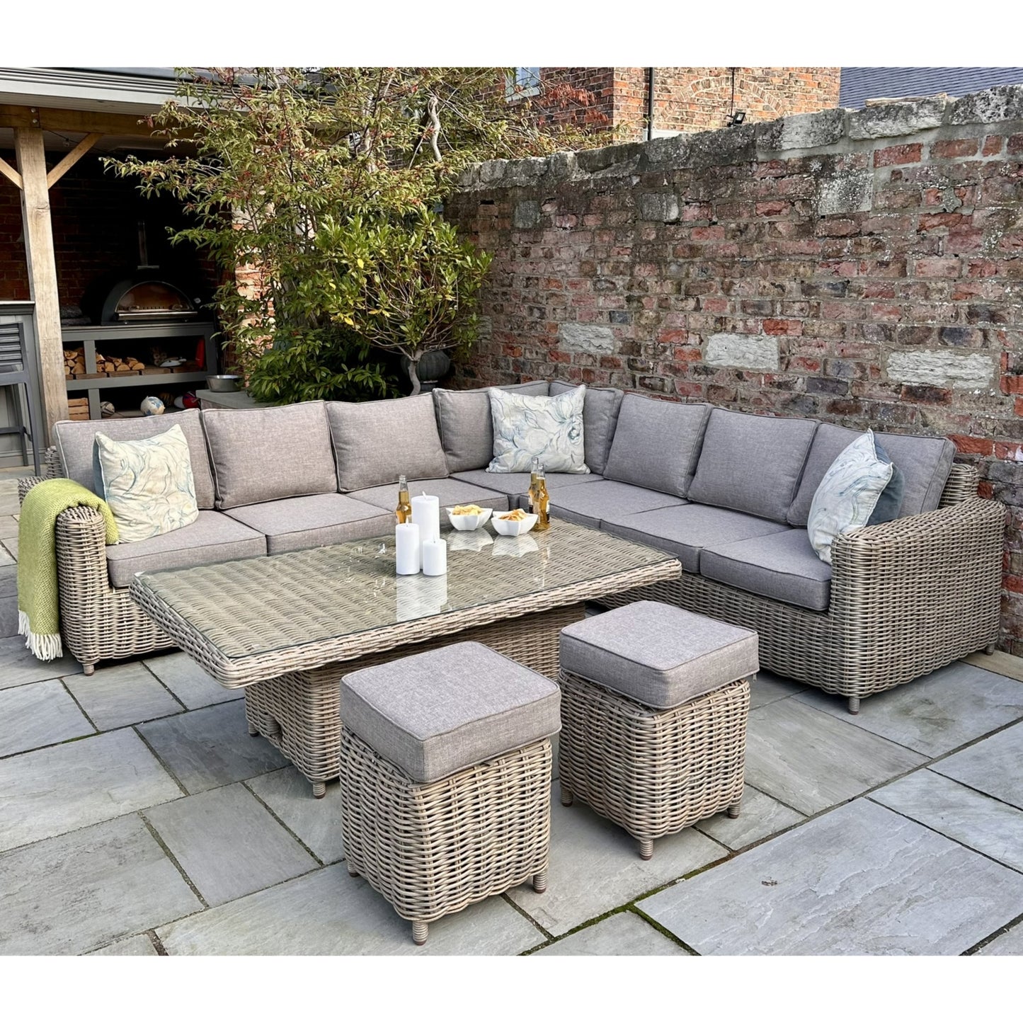Stylish outdoor corner set featuring grey cushions, a glass-top riser table, and two matching stools, surrounded by stone tiles.
