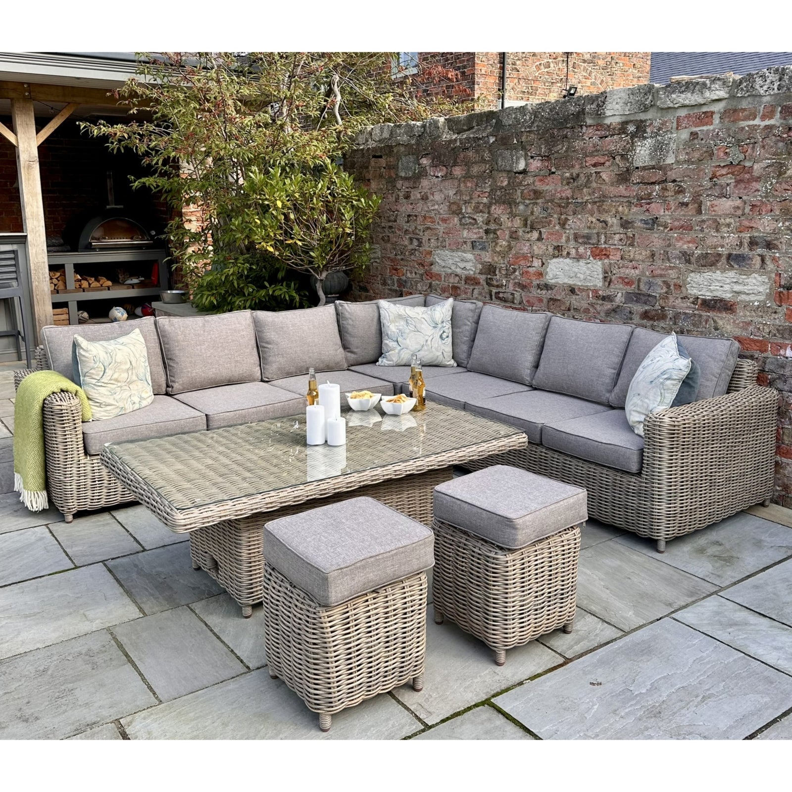Stylish outdoor corner set featuring grey cushions, a glass-top riser table, and two matching stools, surrounded by stone tiles.