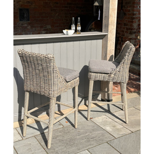 Stylish outdoor bar stools with woven light gray rattan, featuring soft gray cushions, set against a rustic bar backdrop.