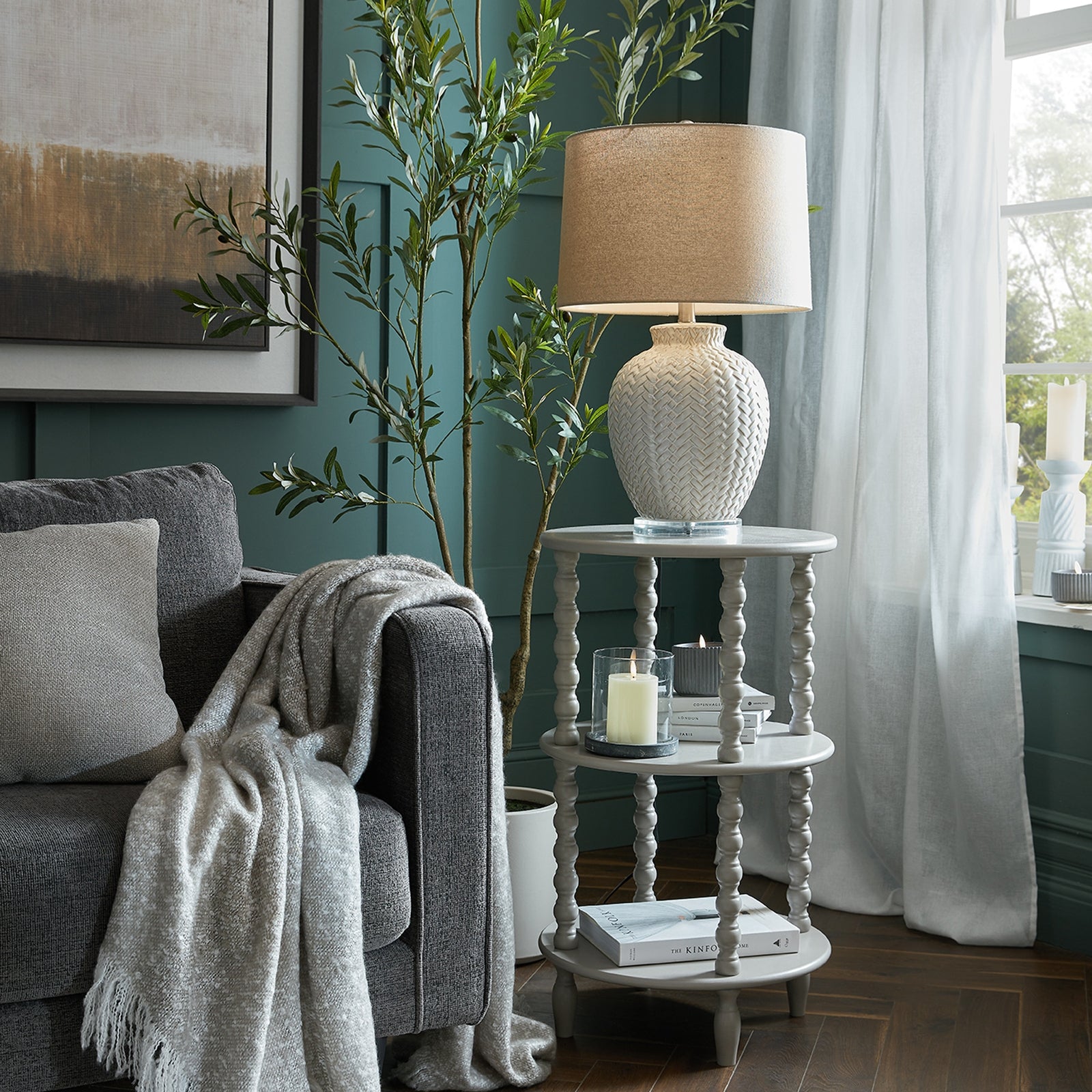 Tall round side table in soft gray with elegant turned legs, styled with a textured lamp, candles, and decorative books.