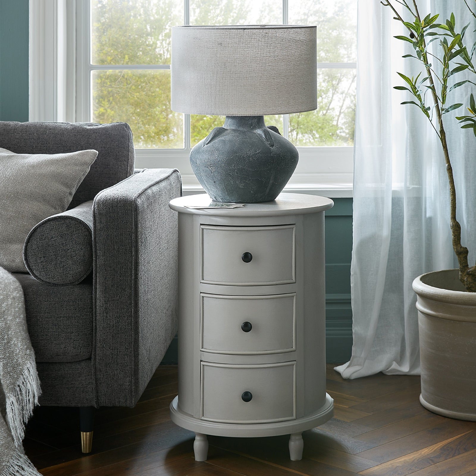 Sophisticated round side table in light gray, featuring three drawers and topped with a textured, stone-like lamp, beside a cozy couch.