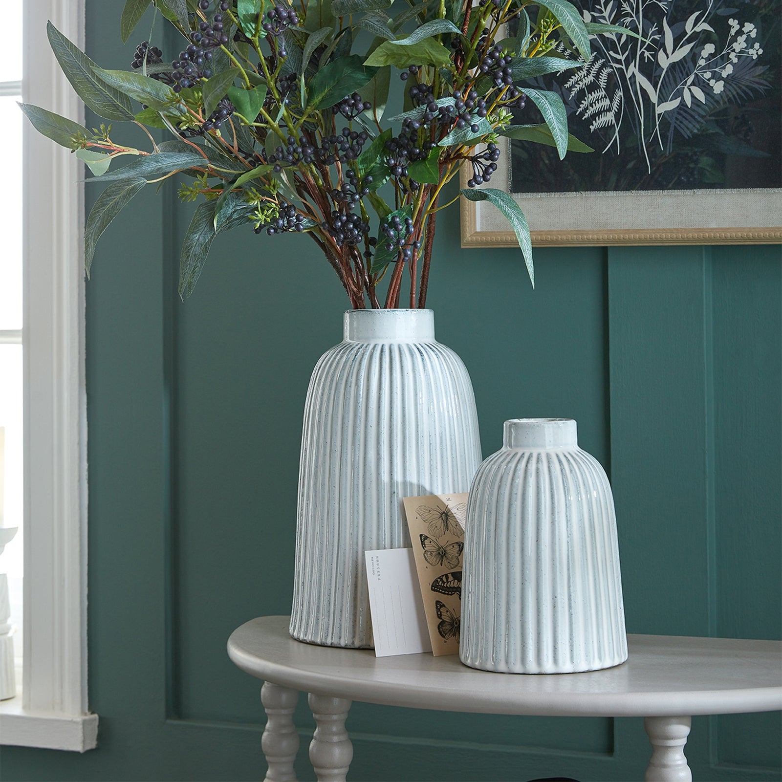 White medium bell vase with a ribbed design, elegantly displaying greenery, set on a round table against a teal wall.