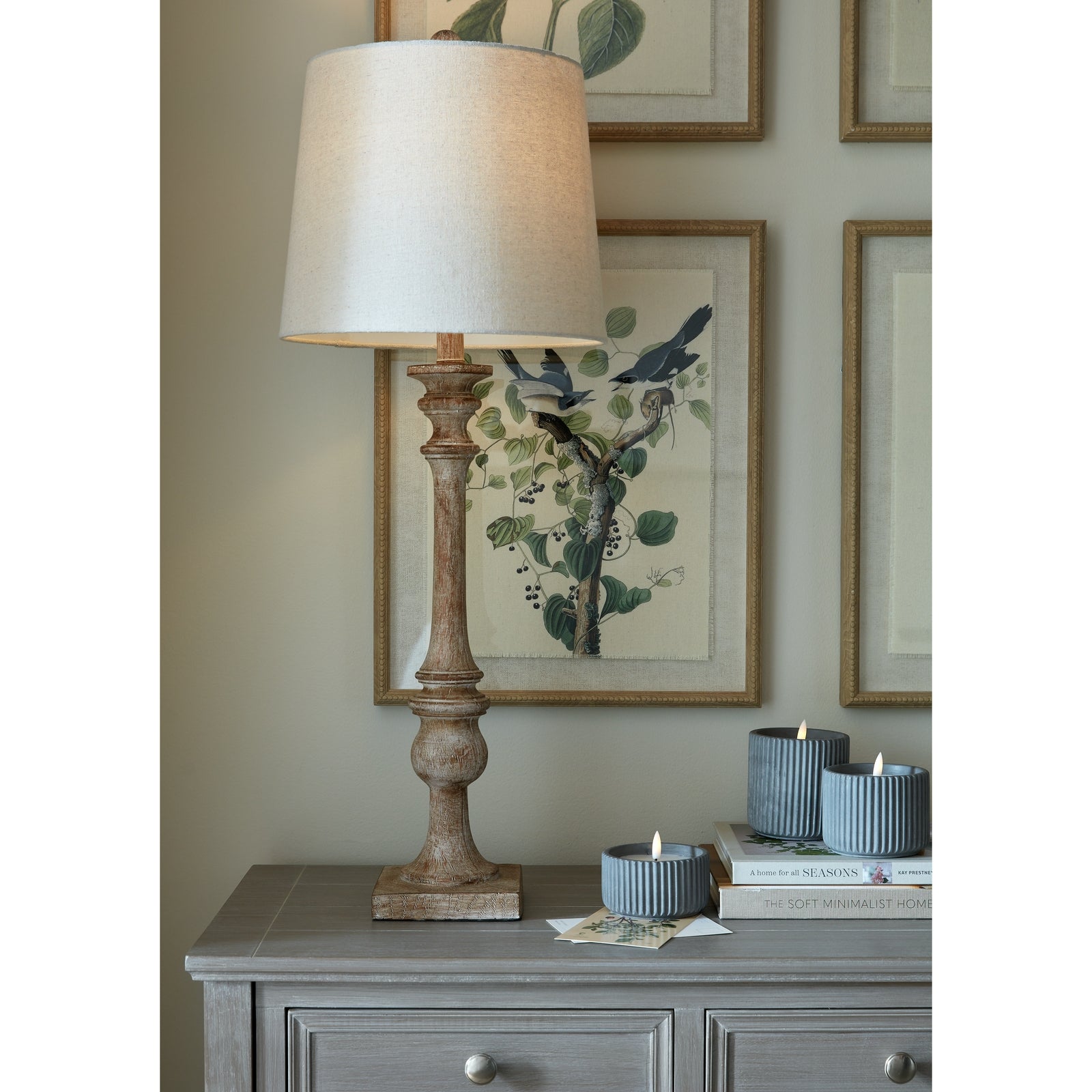 Tall candlestick table lamp with a textured metal base and crisp linen shade, alongside decorative candles and framed botanical art.